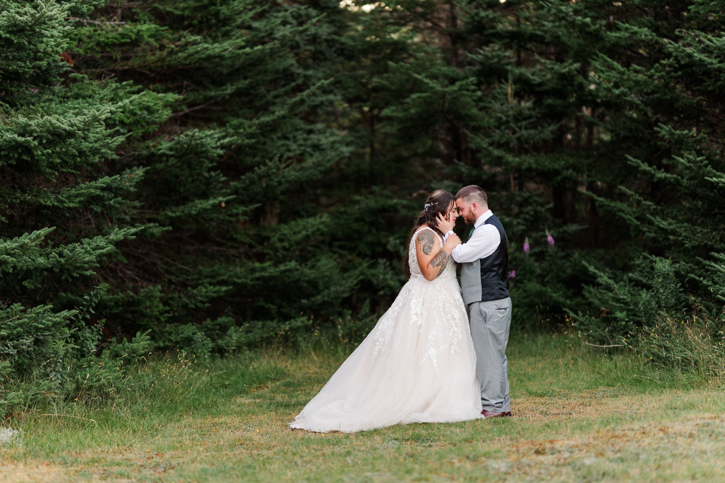 A couple in wedding attire share an intimate moment in front of an evergreen forest backdrop.