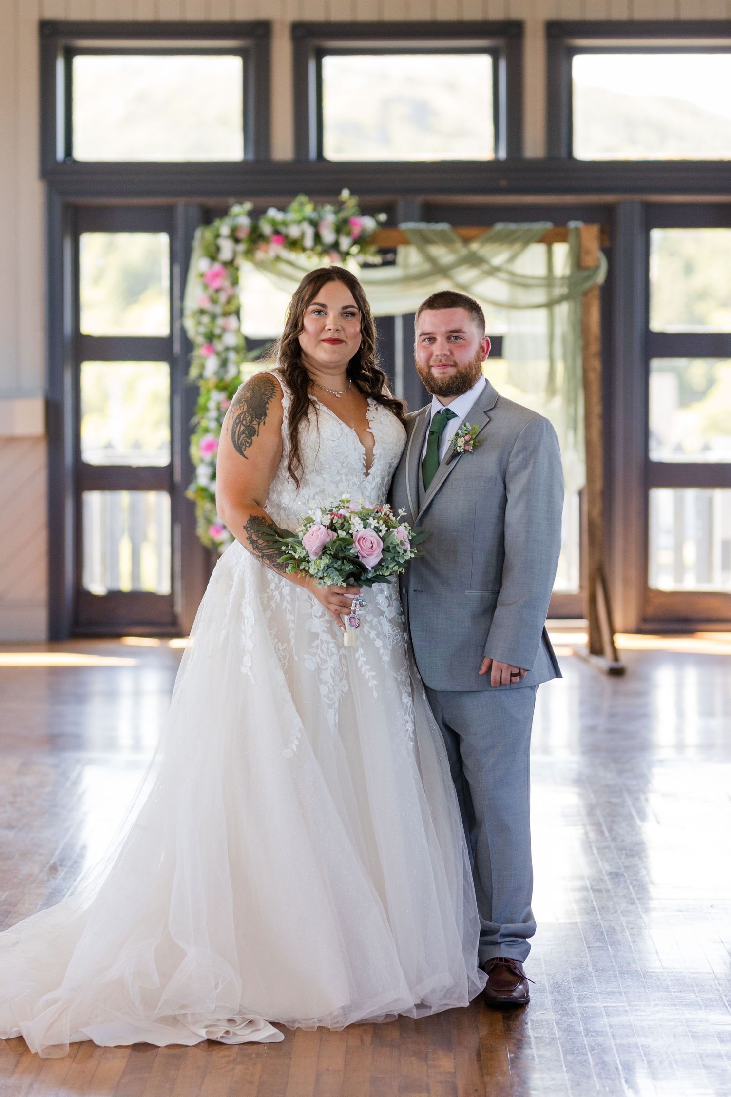 Newlywed couple stands in elegant venue with dark wood floors and windows, holding purple and white floral bouquet.