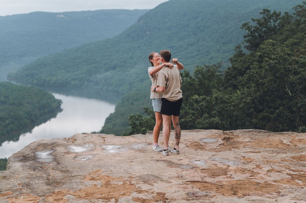 Two people embrace while standing on a rocky overlook above a winding river valley with misty mountains in the background.