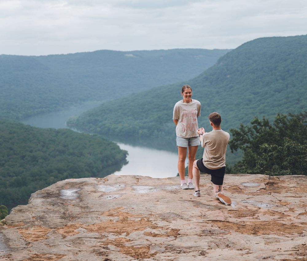 Two people stand on a rocky cliff overlooking a winding river valley with lush green mountains in the background.
