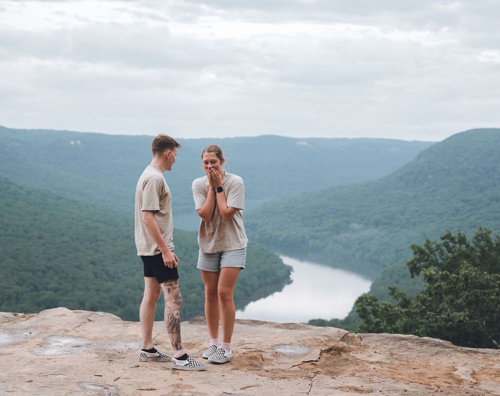 Couple sharing a conversation while standing on a scenic mountain overlook above a winding river and forested valley.