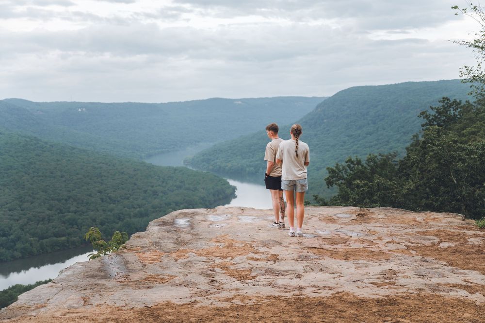 A couple stands on a rocky cliff overlooking a scenic mountain valley with a winding river below on a cloudy day.