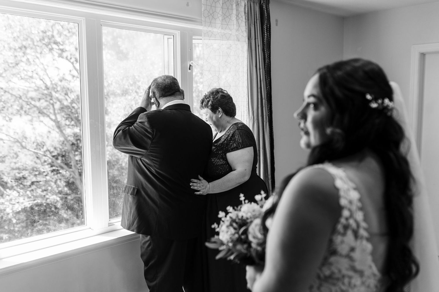 Emotional wedding moment captured in black and white as family members embrace by window while bride holds bouquet.