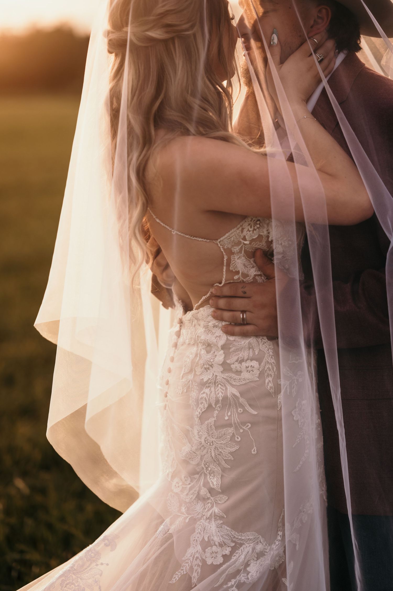 Bride and groom embrace outdoors at golden hour with delicate lace gown and flowing veil details.