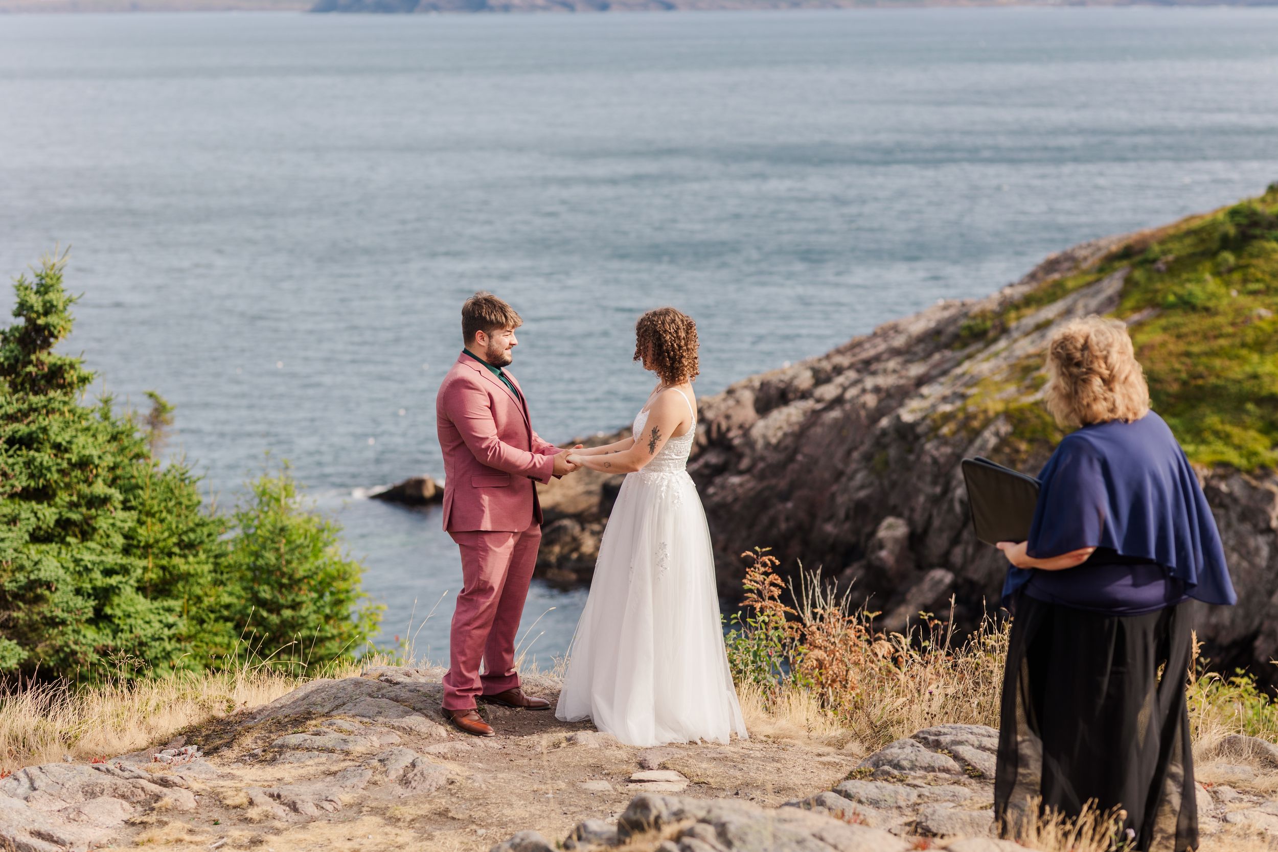 A couple stands on a scenic cliffside overlooking the ocean during a summer outdoor wedding celebration.