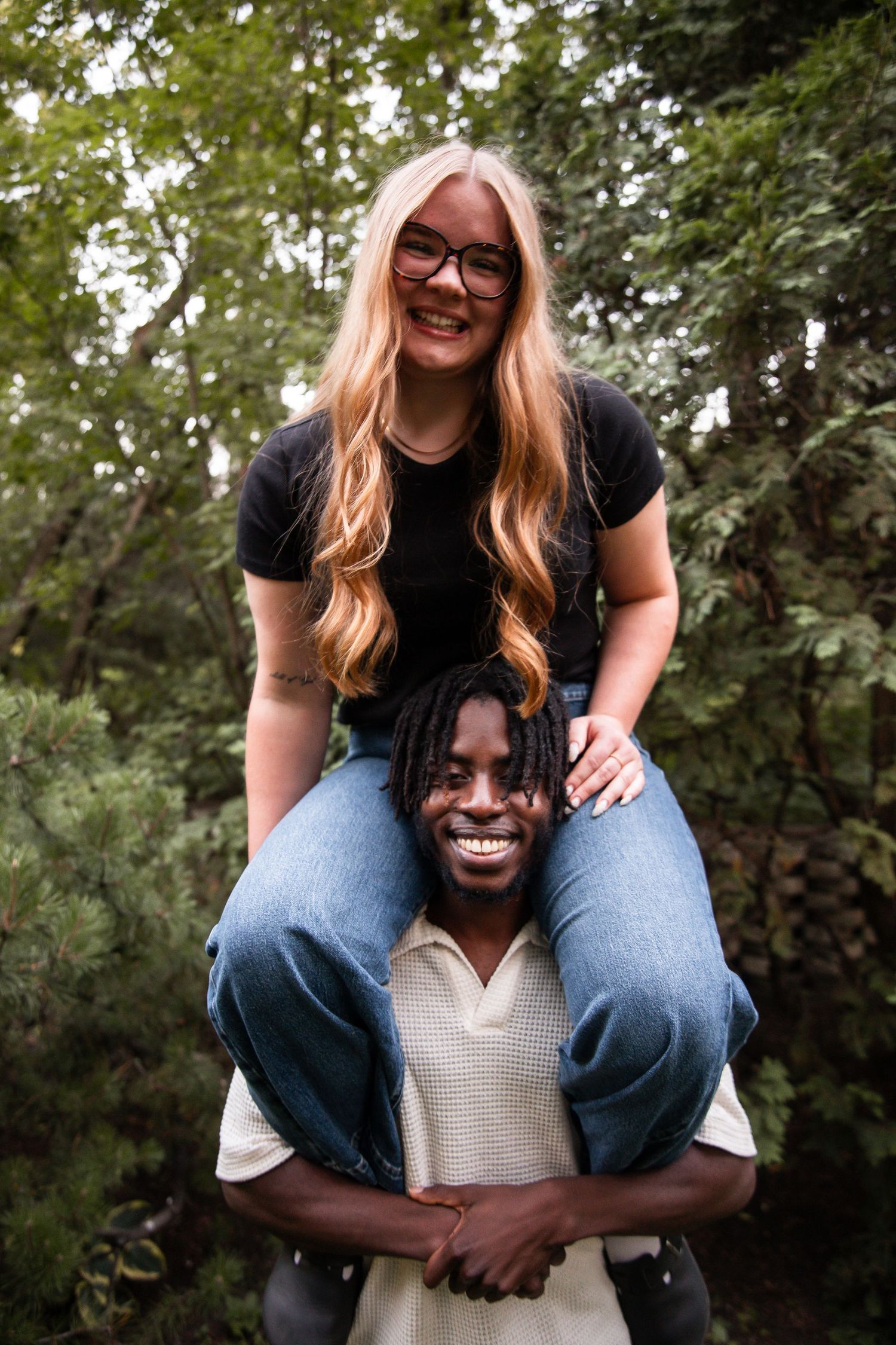A smiling woman with glasses sits on a man's shoulders outdoors, both laughing in a lush green forest setting.