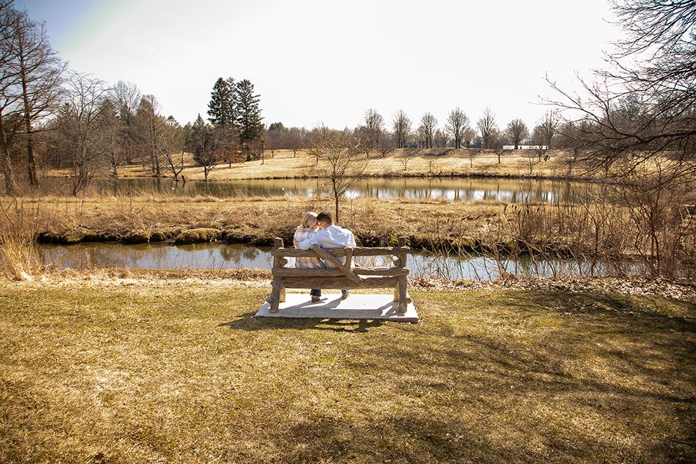 A couple sits on a wooden bench overlooking a scenic pond surrounded by dormant winter grass and bare trees.