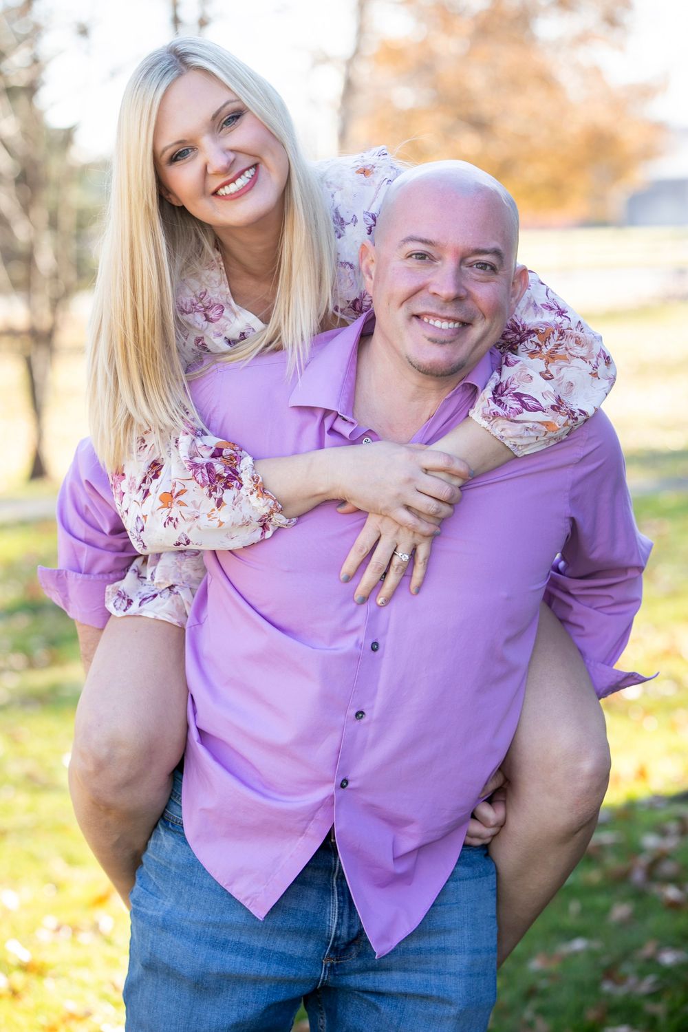 Couple in matching purple tops and jeans posing playfully outdoors with autumn foliage in background.