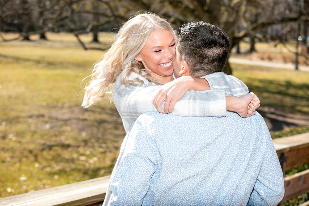 Person in light blue sweater laughs joyfully outdoors in autumn setting with trees in background.