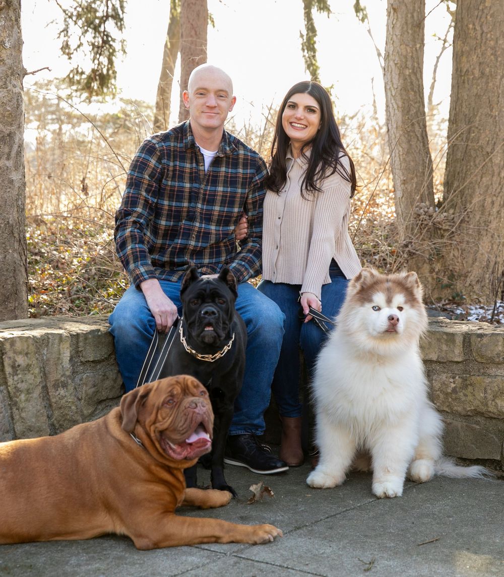 A couple sits with their three dogs, including a Dogue de Bordeaux, black Lab and fluffy white Samoyed, on stone steps outdoors.