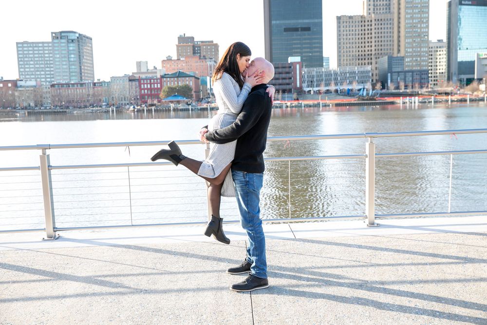 Couple embraces and shares a romantic kiss on a deck overlooking a city waterfront and skyscrapers.