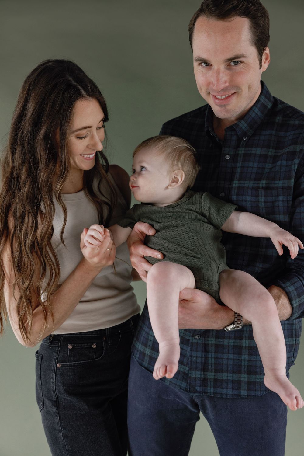 A family sharing a tender moment together with their baby in casual attire against a simple green background.