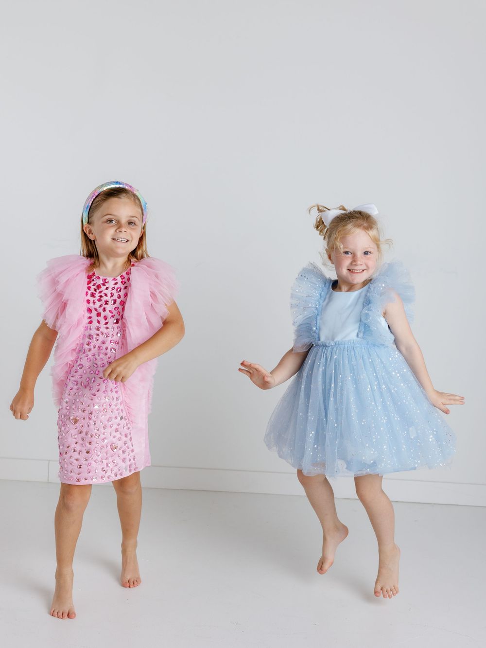 Two children in tulle dresses, one pink and one blue, dancing and playing barefoot against a white backdrop.