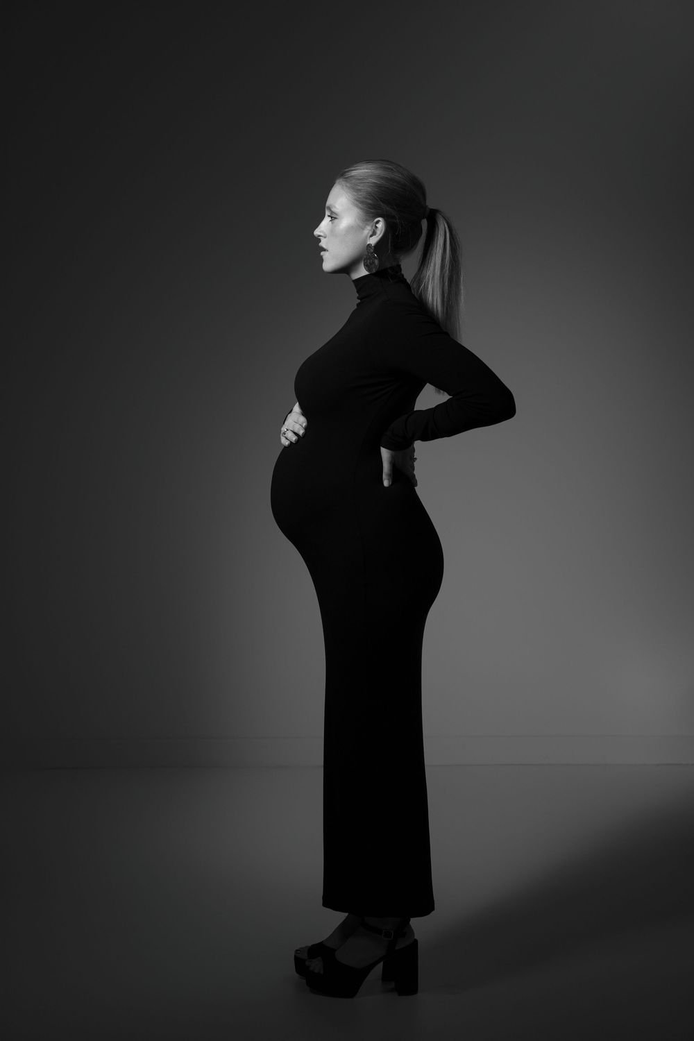 Black and white artistic maternity portrait featuring a silhouette in a long fitted dress against a dark background.