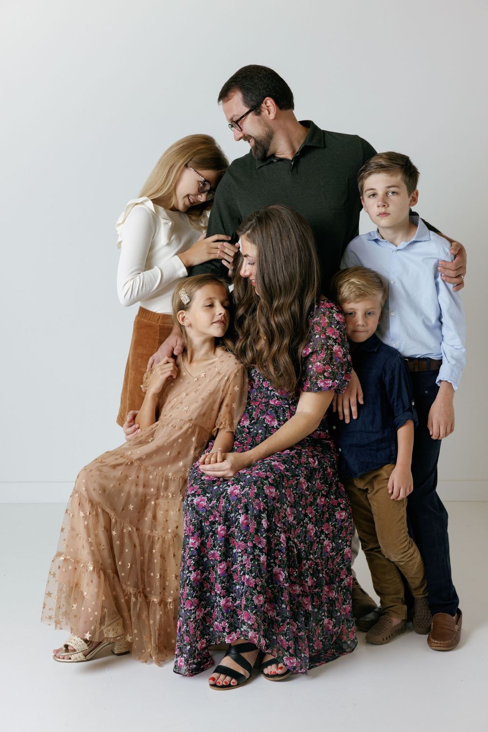 Family portrait in coordinated outfits with floral dresses and casual attire against white backdrop, gathered close together.