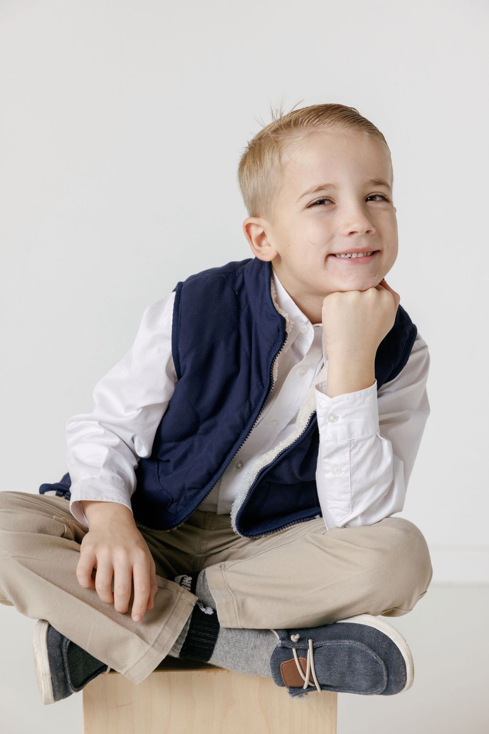 A young boy in a navy vest and white shirt sits cross-legged while smiling and resting his chin on his hand.