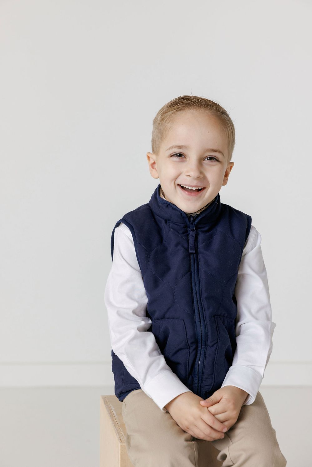 Cheerful young child wearing a navy vest and white shirt sits casually against white background.
