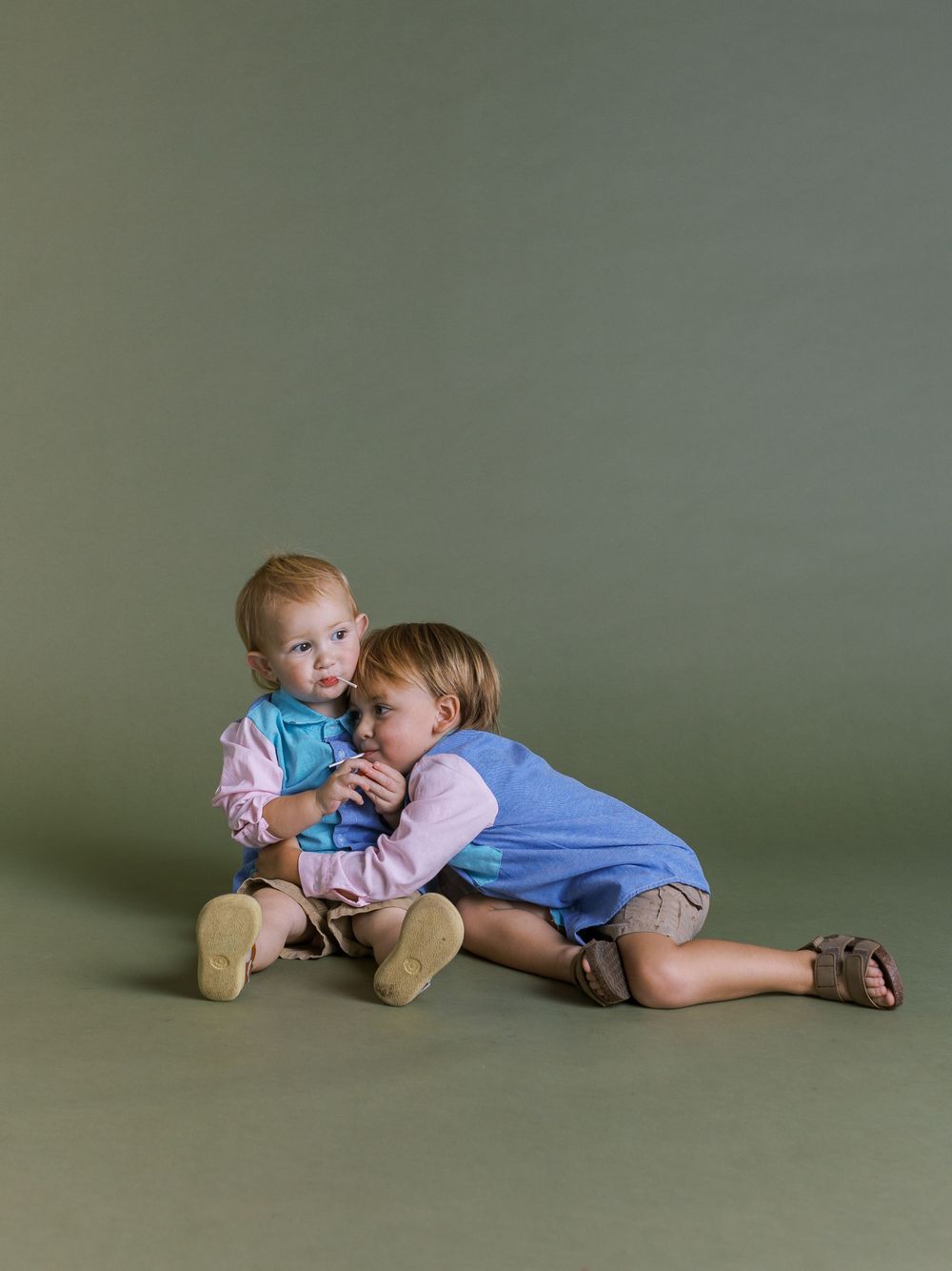 Two small children sit together wearing light blue and pink outfits against a sage green studio backdrop.