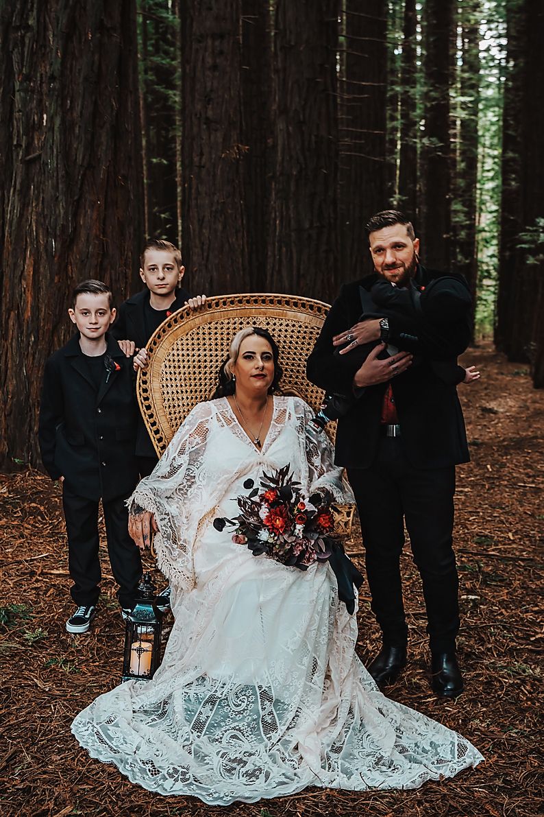 Wedding party poses in redwood forest with couple seated on vintage wicker chair among tall trees.