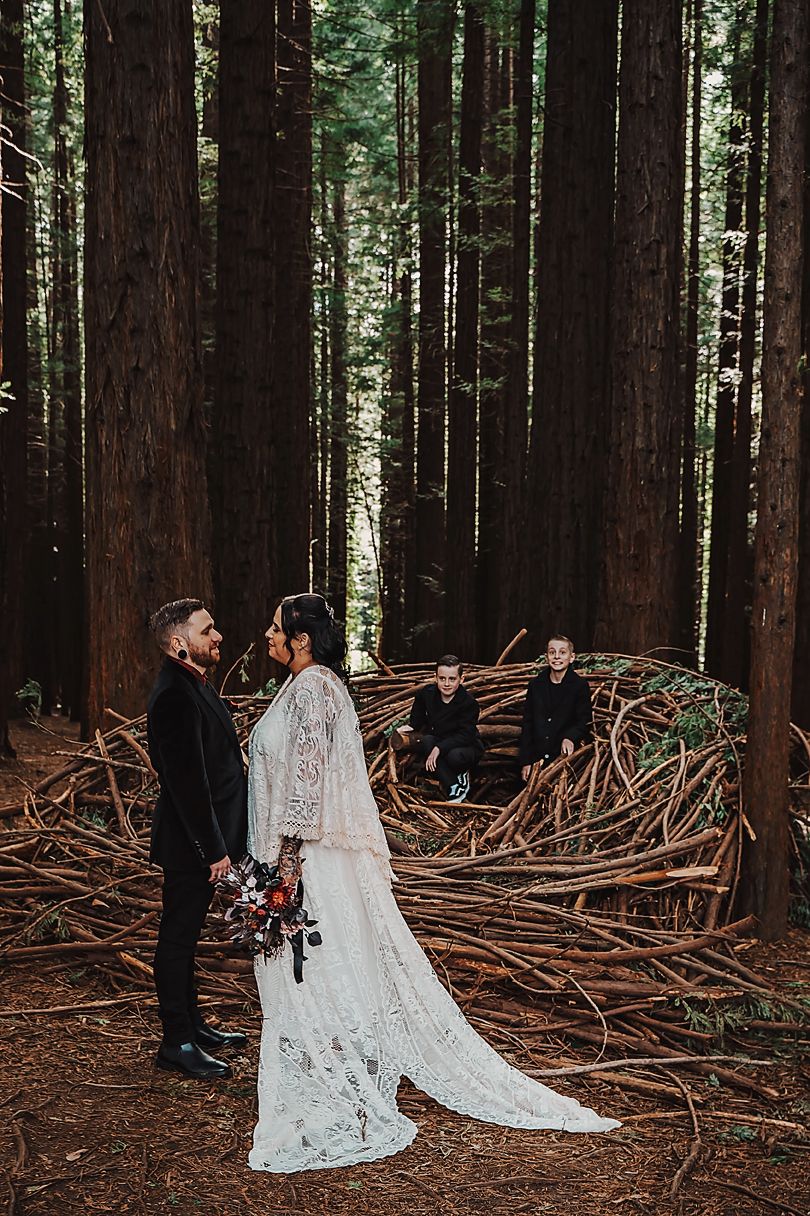 A couple celebrates their wedding ceremony in a majestic redwood forest surrounded by towering trees and fallen branches.