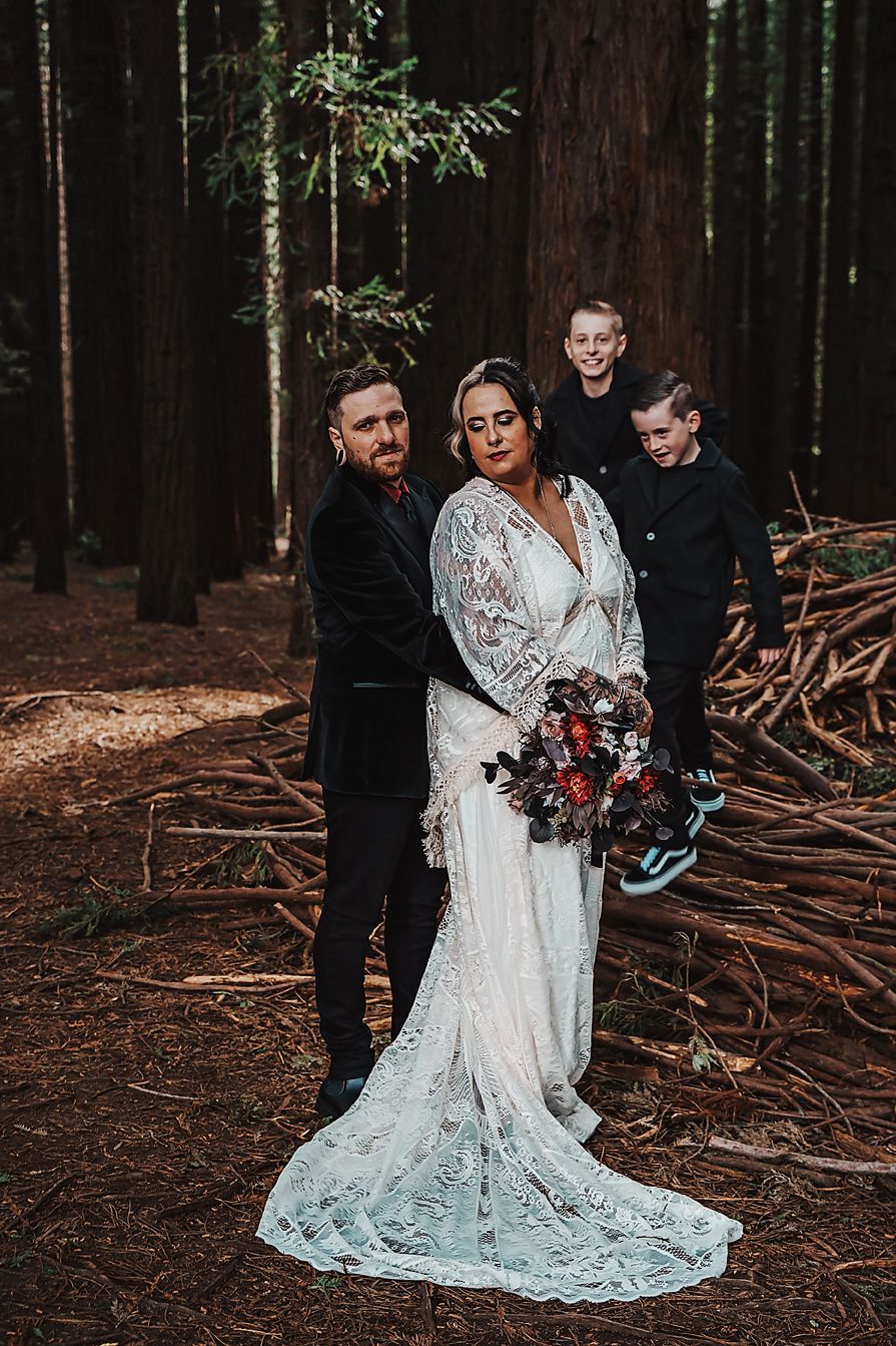 Wedding group photo in moody forest setting with white lace gown and dark suits against redwood trees.