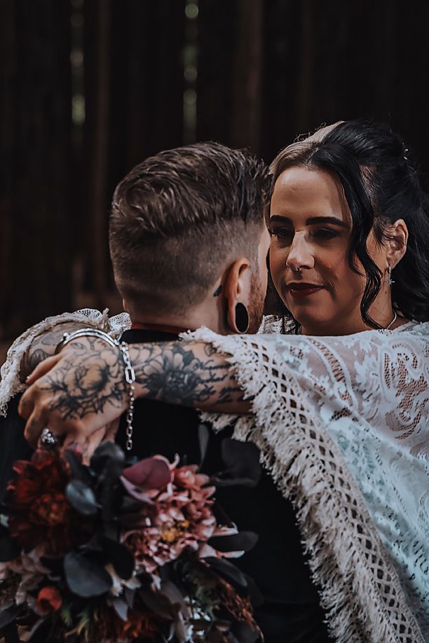 Romantic couple embraces in a dark forest setting with lace wedding dress details and moody fall-toned bouquet.