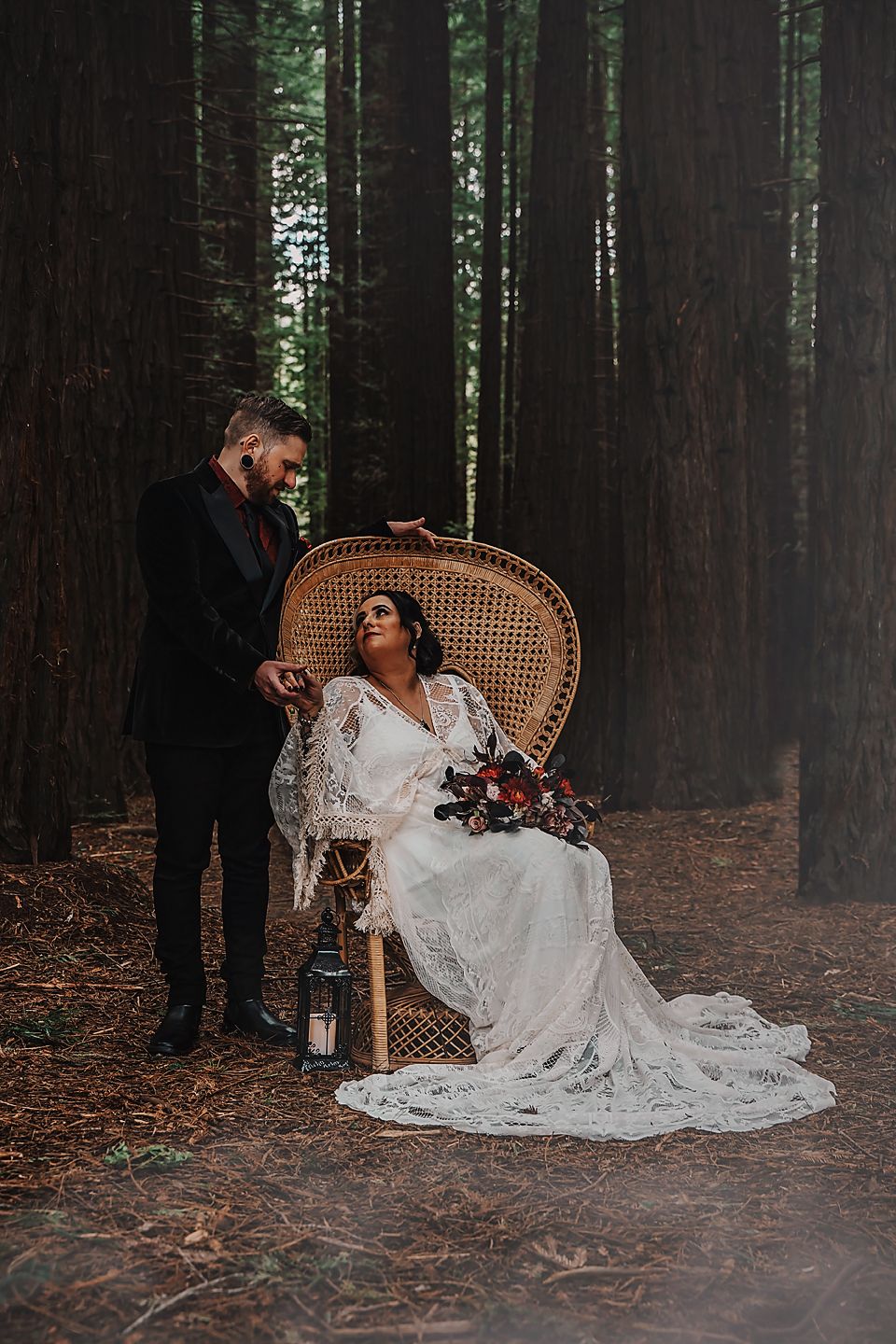 A romantic forest wedding portrait featuring someone in a flowing lace wedding dress seated on a rattan peacock chair.