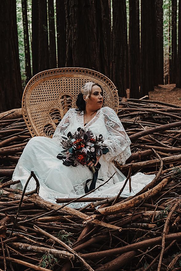 A bride in a white lace dress reclines on a peacock chair surrounded by redwood trees and fallen logs.