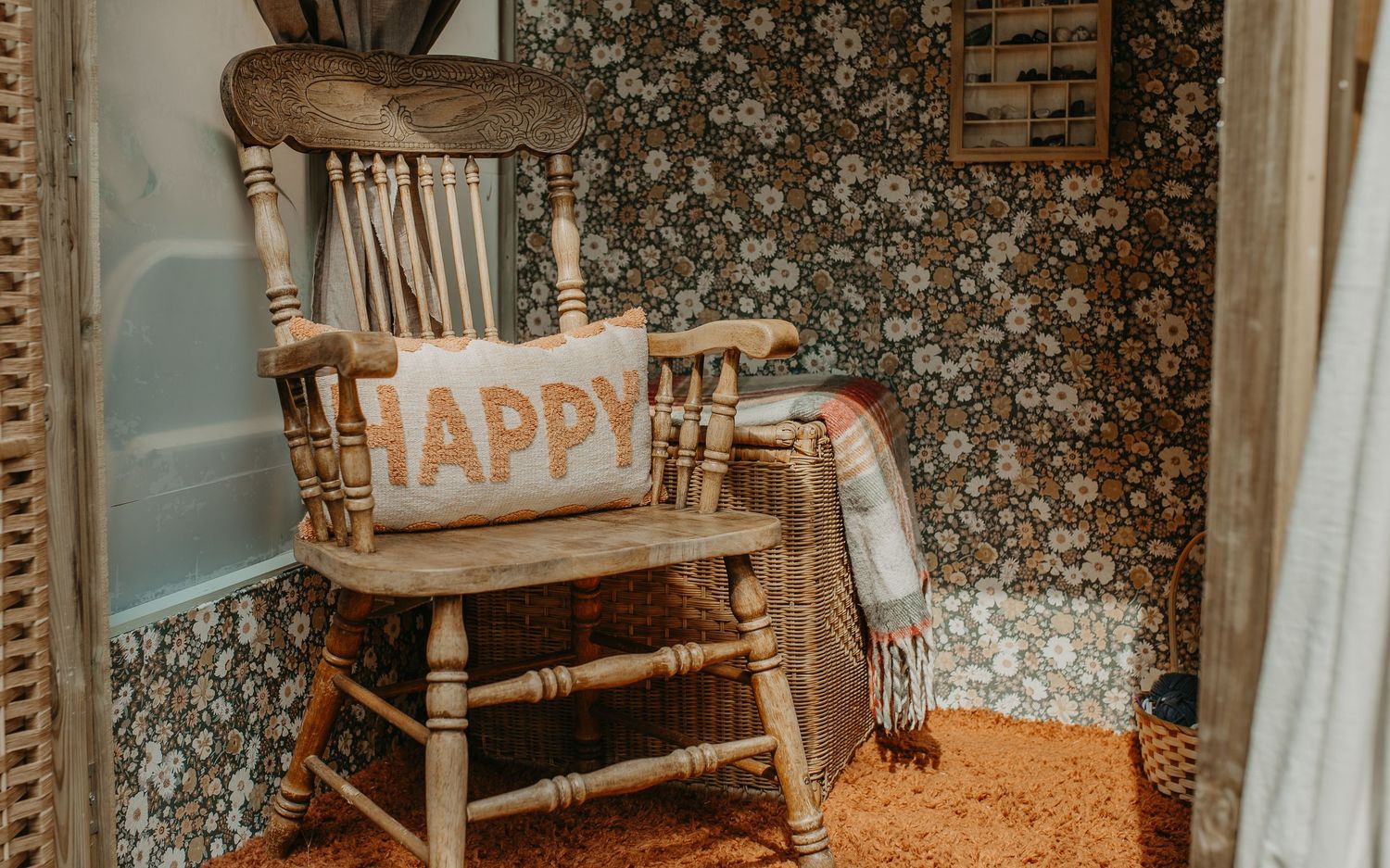 Rustic wooden rocking chair with decorative 'Happy' pillow sits against vintage floral wallpaper in cozy corner.