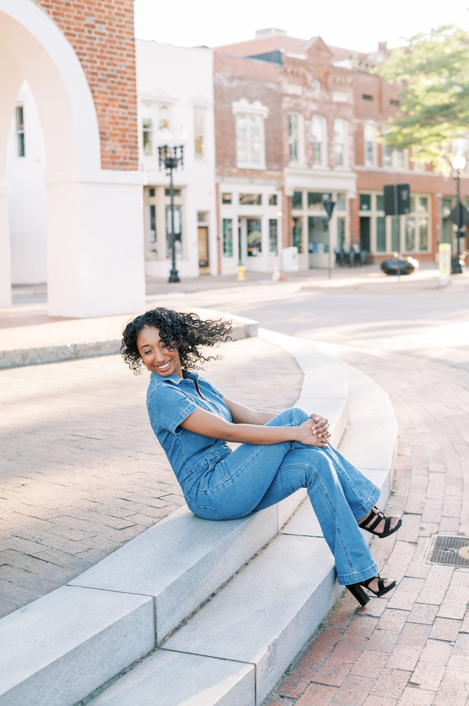 A candid, playful image of a woman wearing a denim jumpsuit sits on concrete steps in downtown Fayetteville.