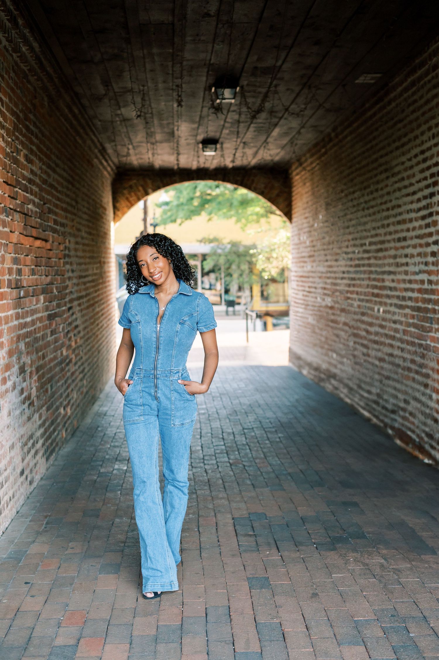 A woman of color posing in a brick tunnel in dowtown Fayetteville NC wearing a denim jumpsuit, creating a progressive movement effect.