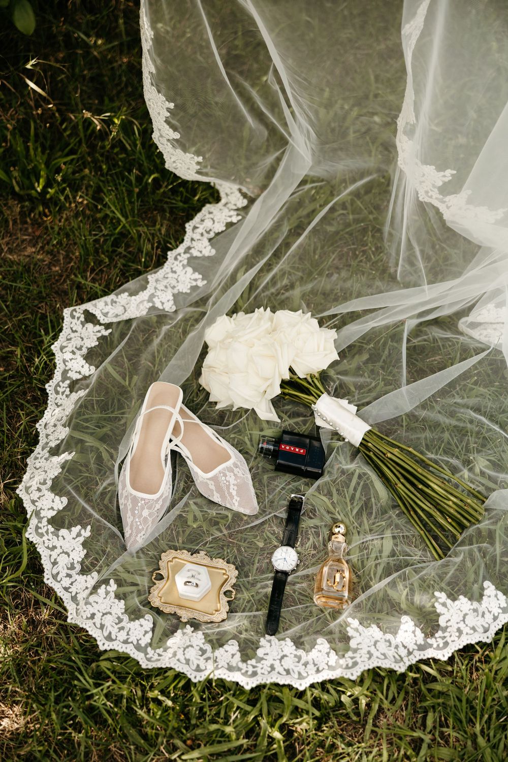 Beautiful bridal accessories including lace veil, perfume bottles, and delicate white shoes arranged on grass.