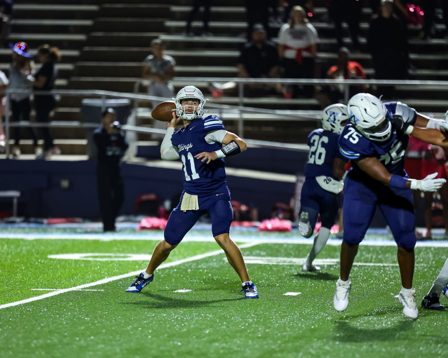 Football player in white and blue uniform steps back to throw pass during nighttime game on turf field.