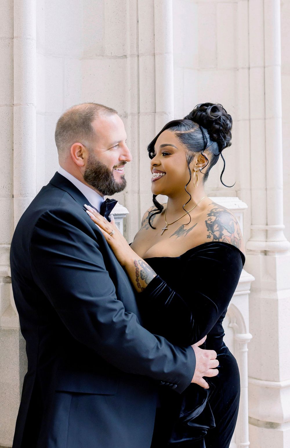 A happy couple laughs together in formal attire against a white stone backdrop.