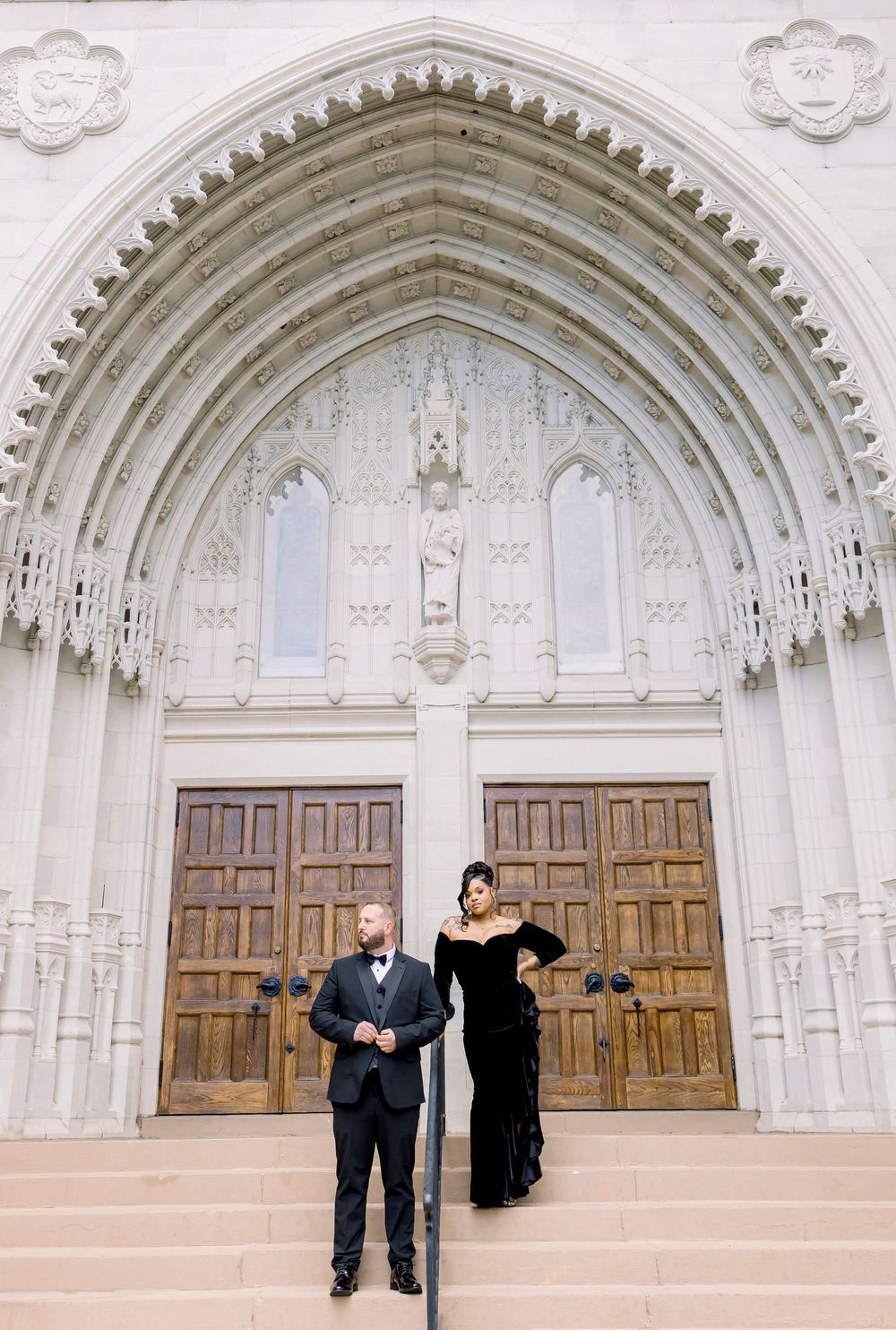 Couple poses elegantly in front of ornate church doors with gothic arched entrance and decorative stonework.