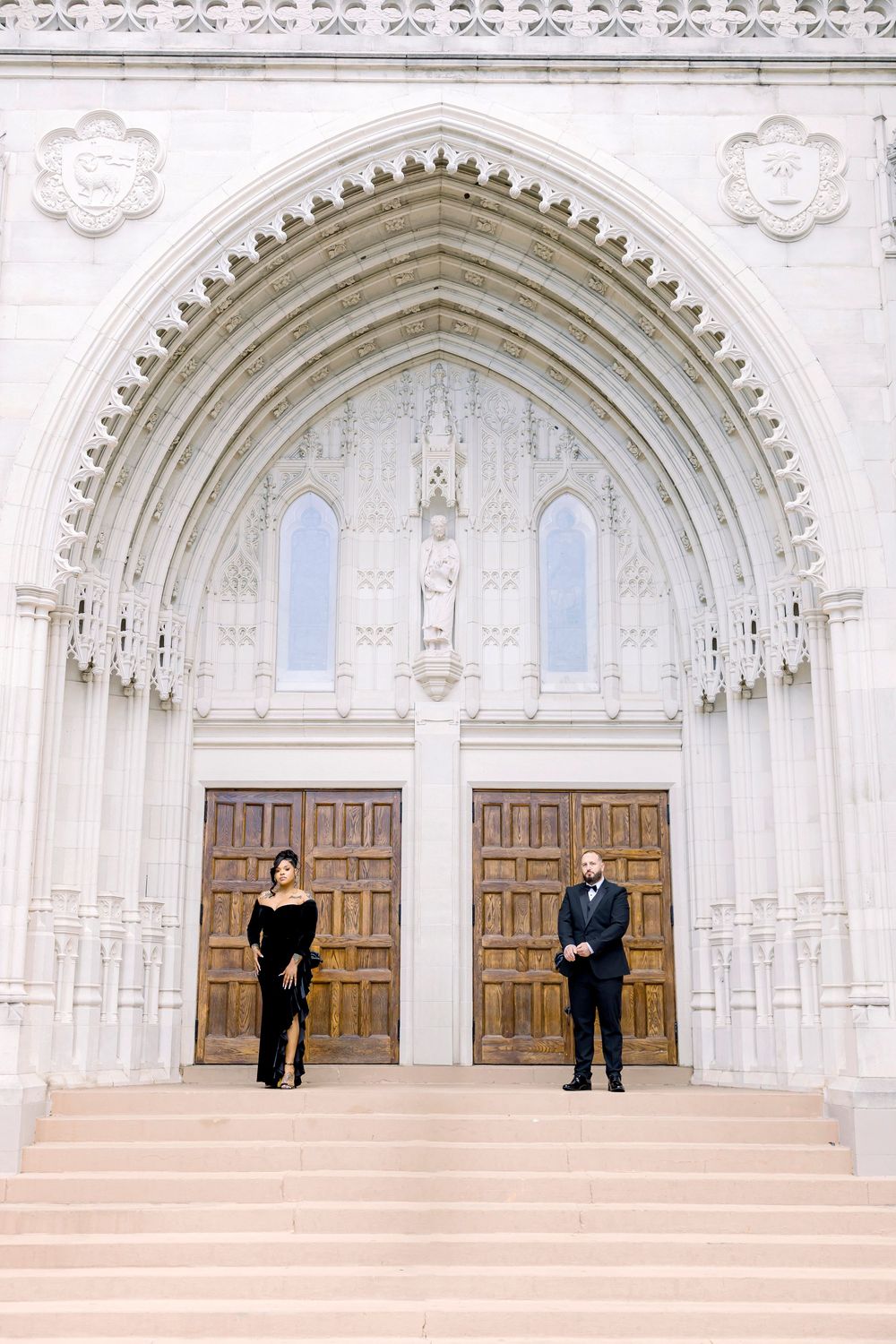 Two people stand in formal attire in front of ornate Gothic church doors with arched architecture.
