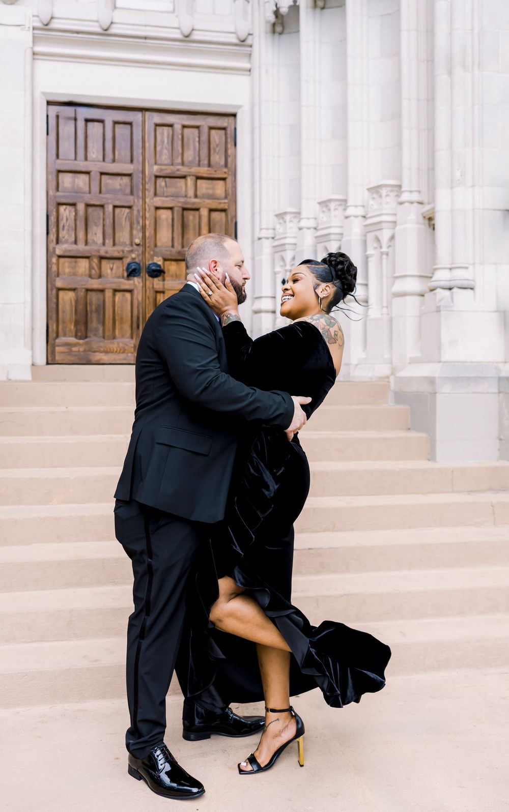 Romantic couple embraces on church steps, with woman in flowing black velvet dress and man in dark suit.