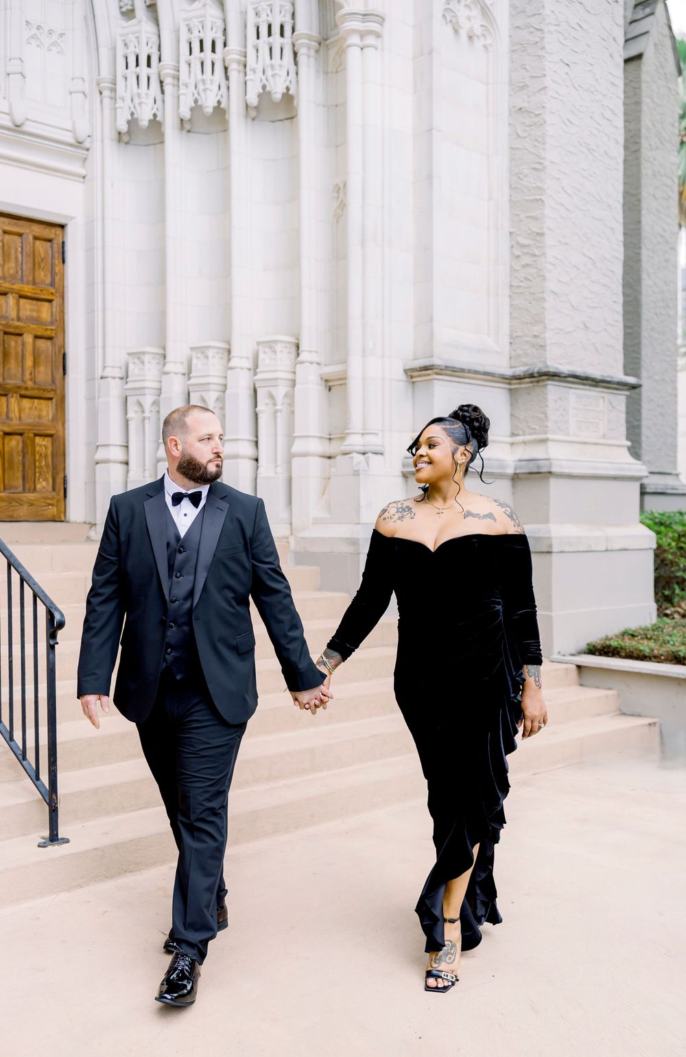 A couple shares a tender moment in formal wear against a white stone church facade.