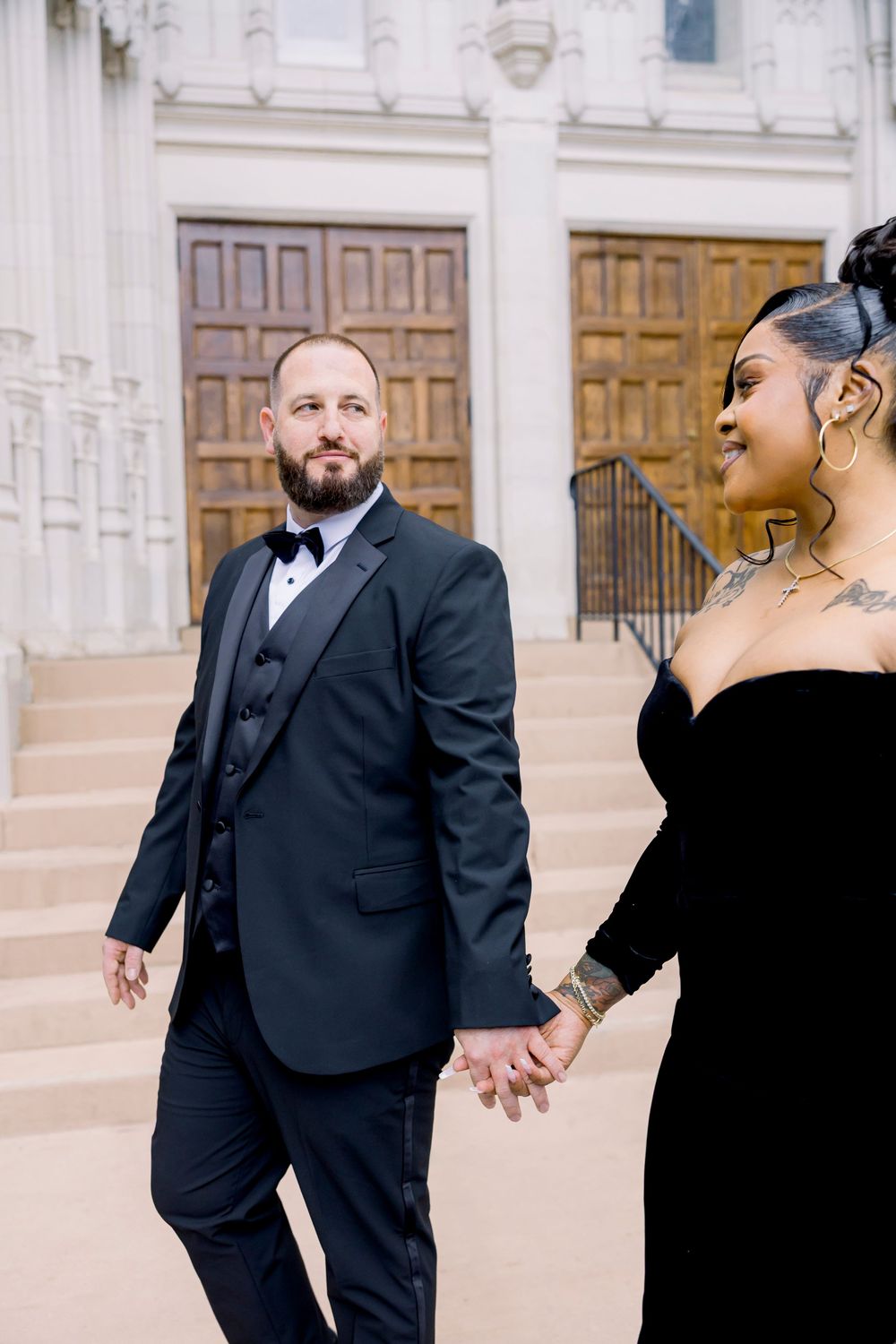 Two people in black formal wear share intimate moments in front of a white building with wooden doors.