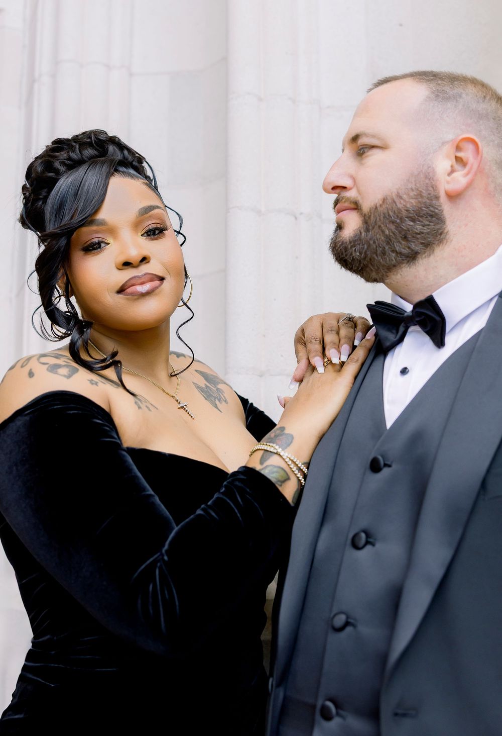Elegantly dressed couple in formal black attire pose together against white columns in romantic photoshoot series.