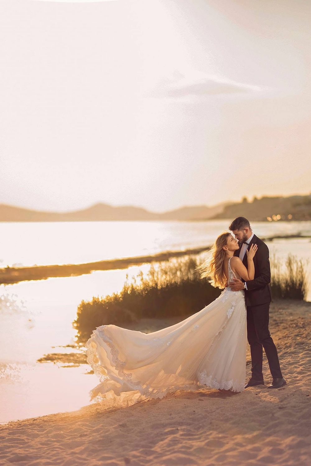 A romantic wedding couple embraces on a sandy beach at sunset with a flowing white dress catching the golden light.