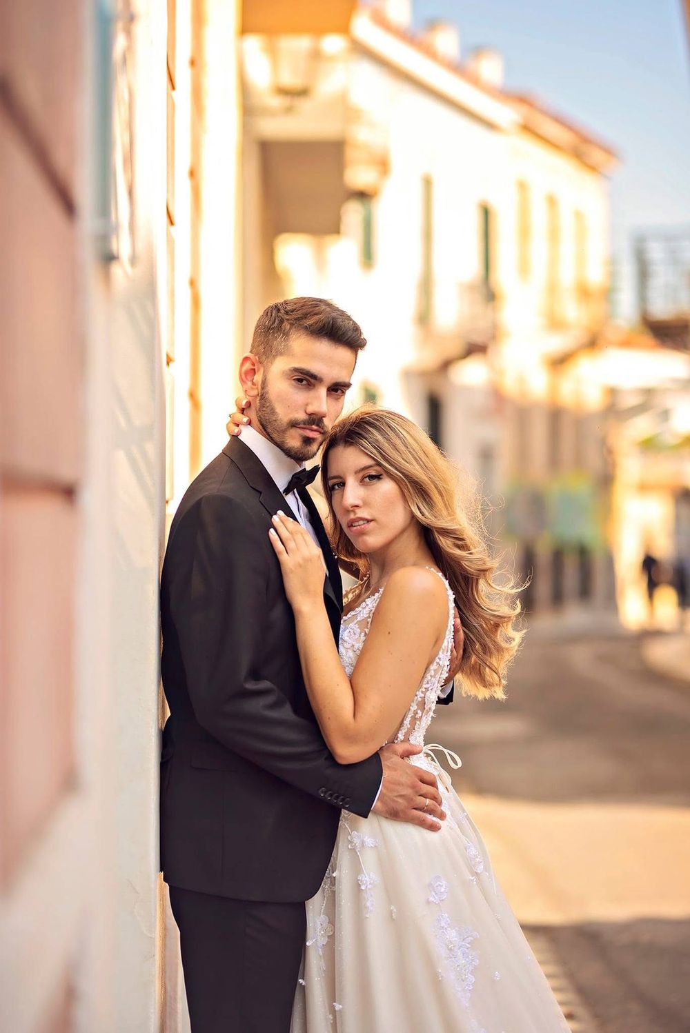 A romantic couple in wedding attire embraces on a sunlit street as golden hour light illuminates the scene.