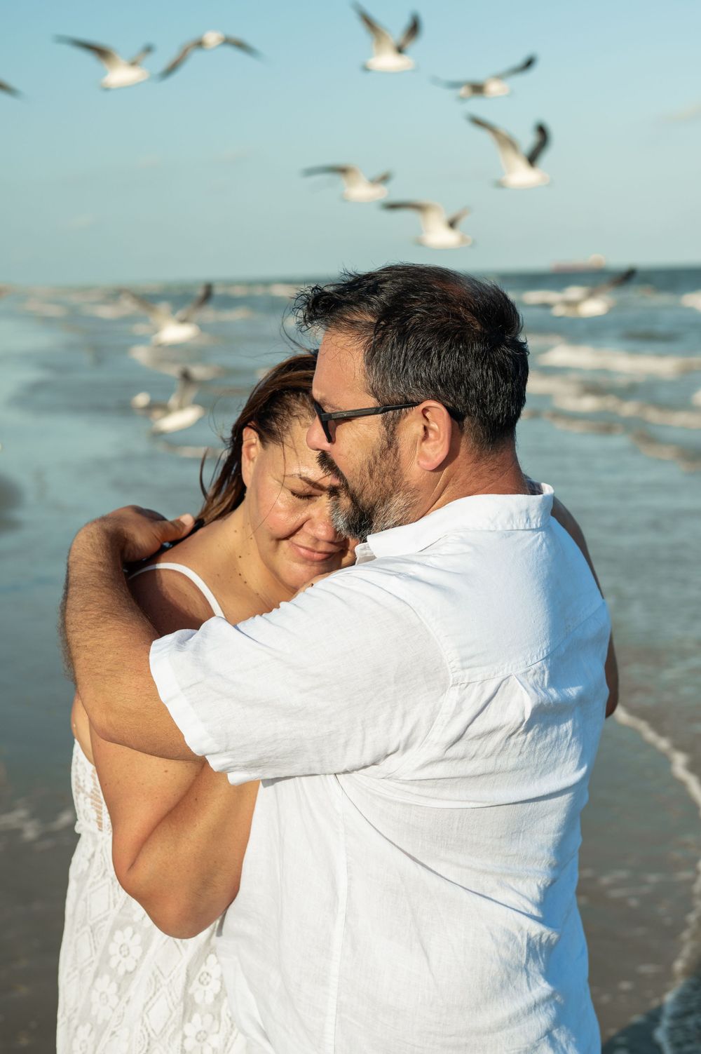 Seagulls soar through the sky as a couple shares a loving embrace on a sunny beach.