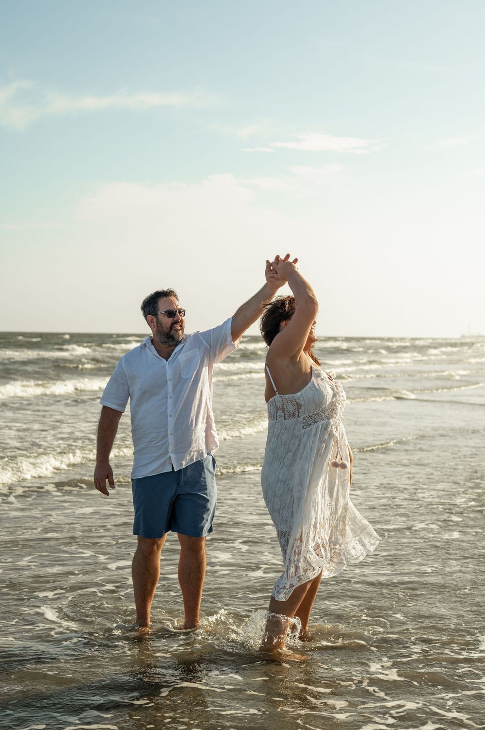 Romantic couple splashing and dancing together in ocean waves at sunset wearing white clothing on the beach.