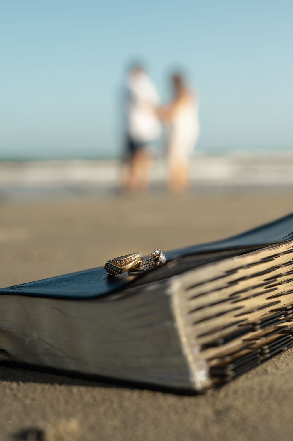 Wedding rings rest on a leather-bound book on sandy beach with blurred couple standing at water's edge in background.