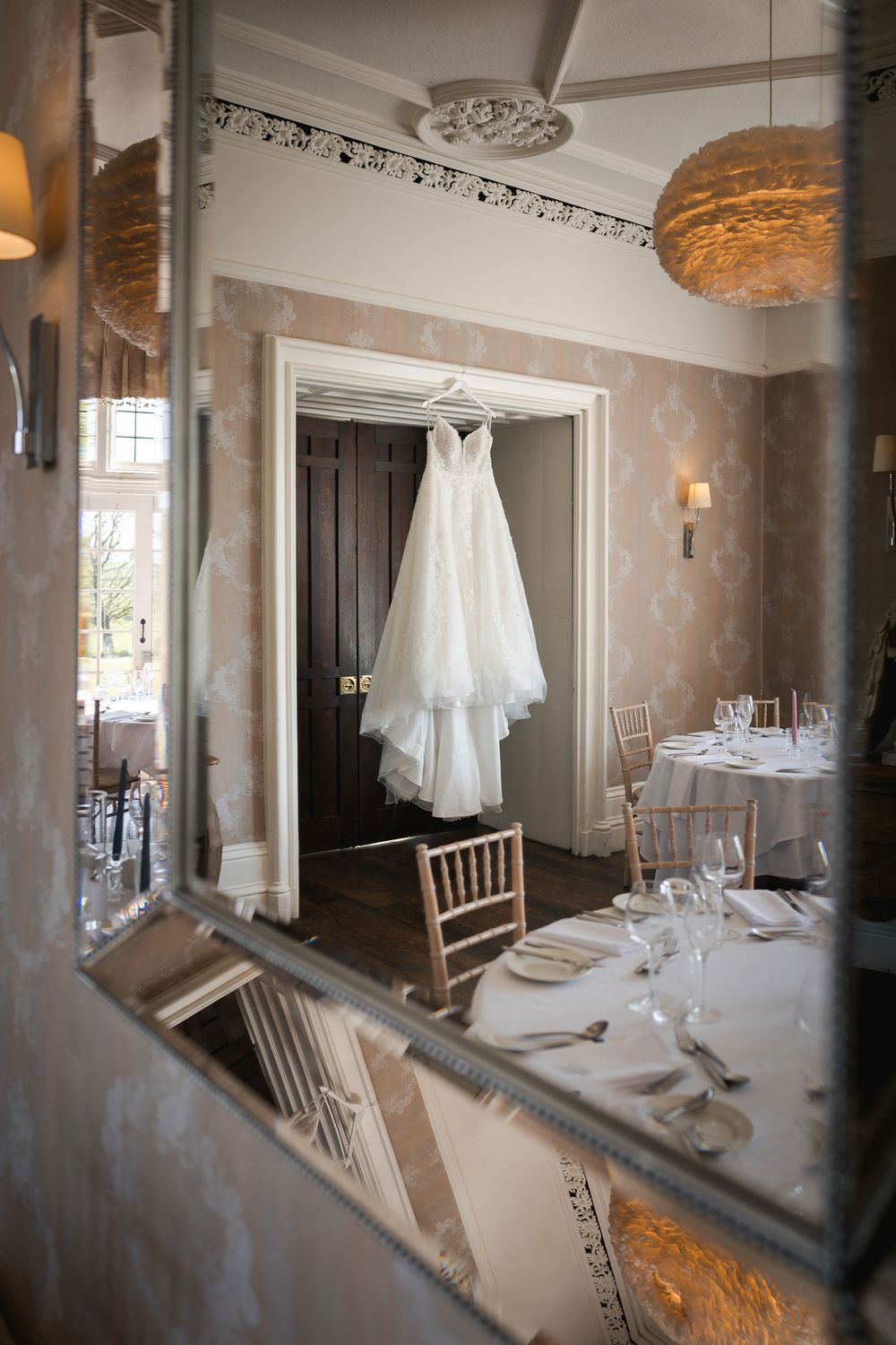 Elegant wedding dress hanging in a sunlit room with ornate decor and place settings.