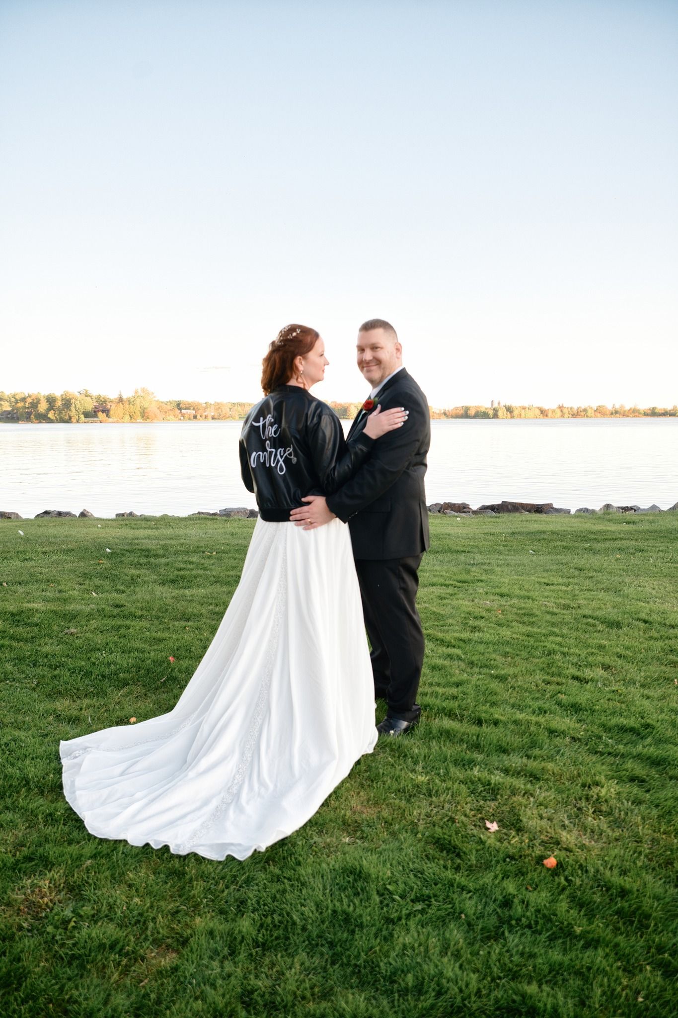 A couple poses on grassy waterfront, bride wearing a white dress with black leather jacket for outdoor wedding photos.