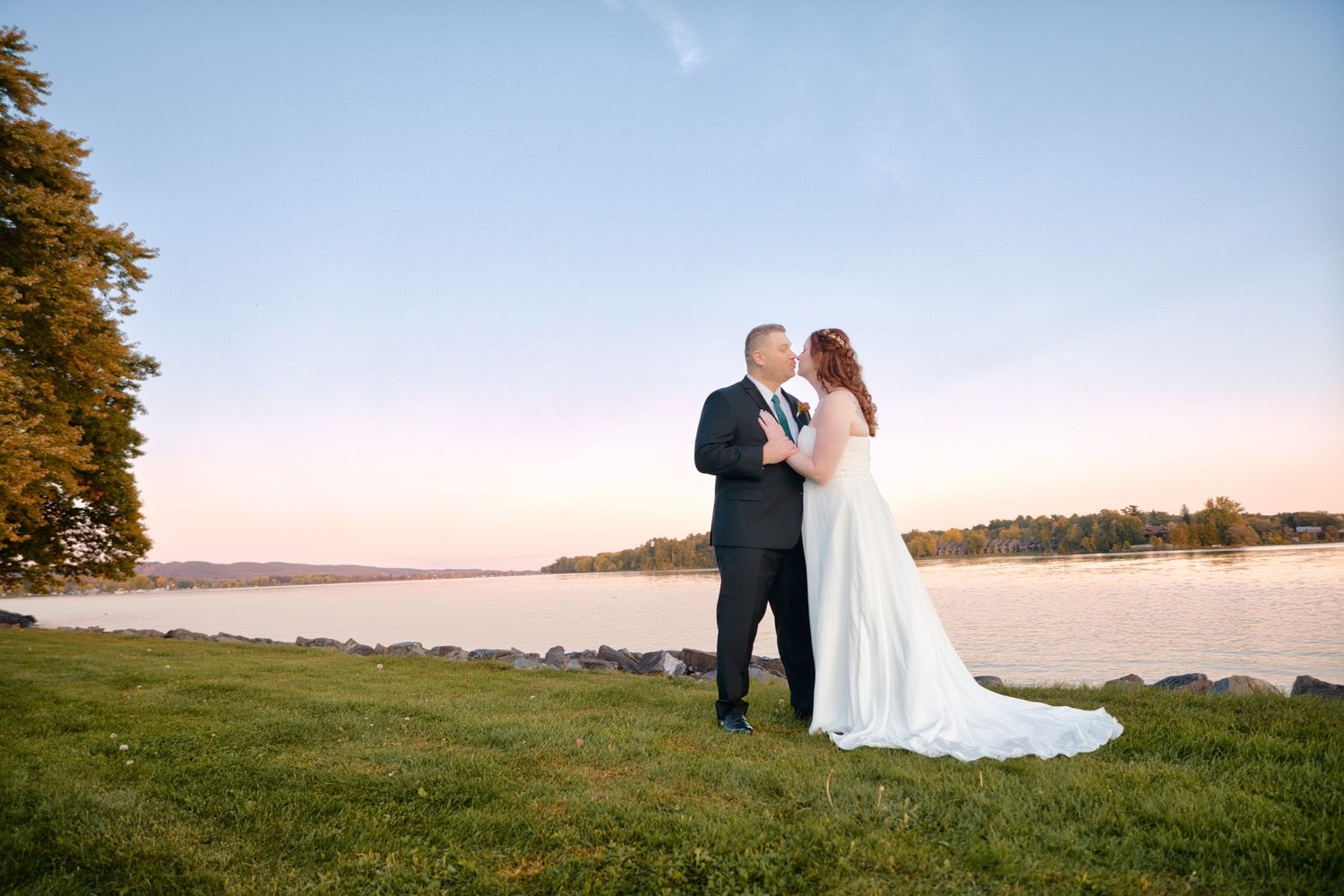 A newlywed couple shares an intimate moment at sunset on a grassy waterfront lawn.