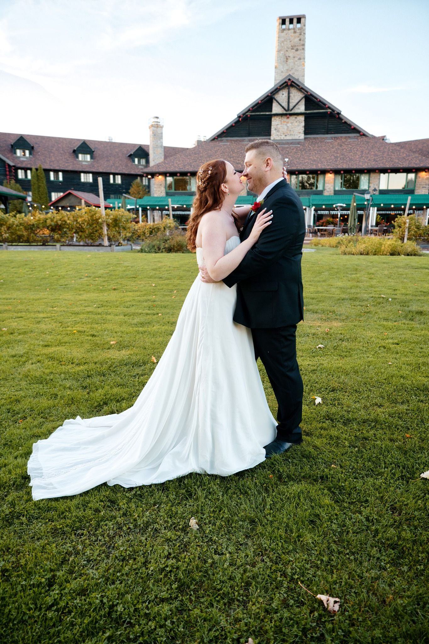 A wedding couple embraces on a green lawn in front of a rustic venue with a distinctive stone chimney.