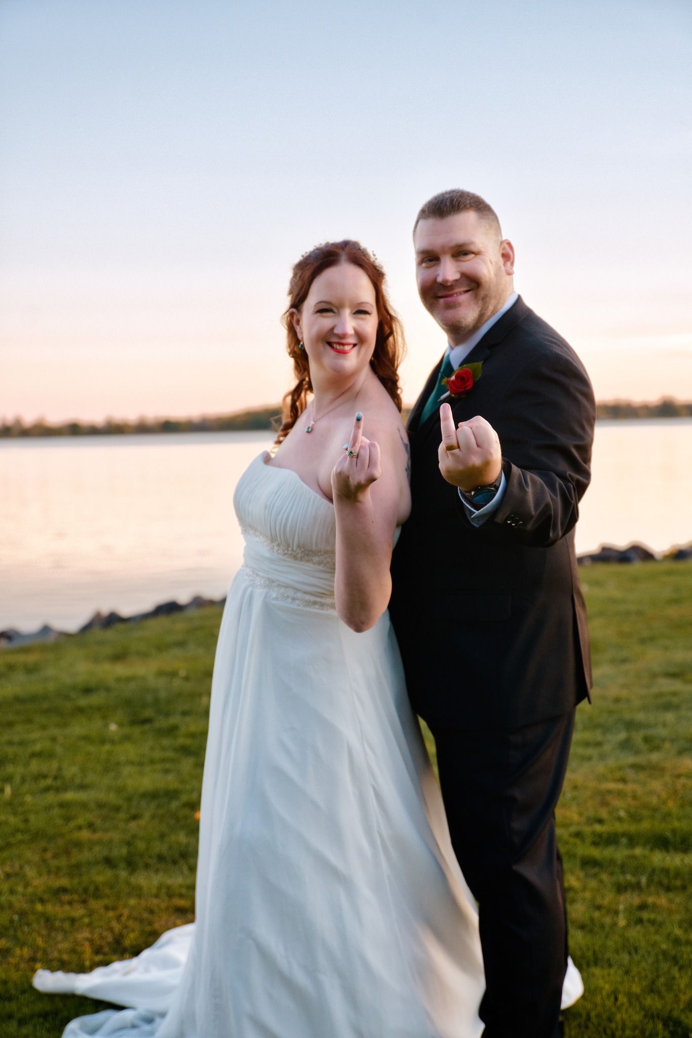 Wedding couple poses playfully outdoors on grass at sunset with blurred waterfront backdrop.